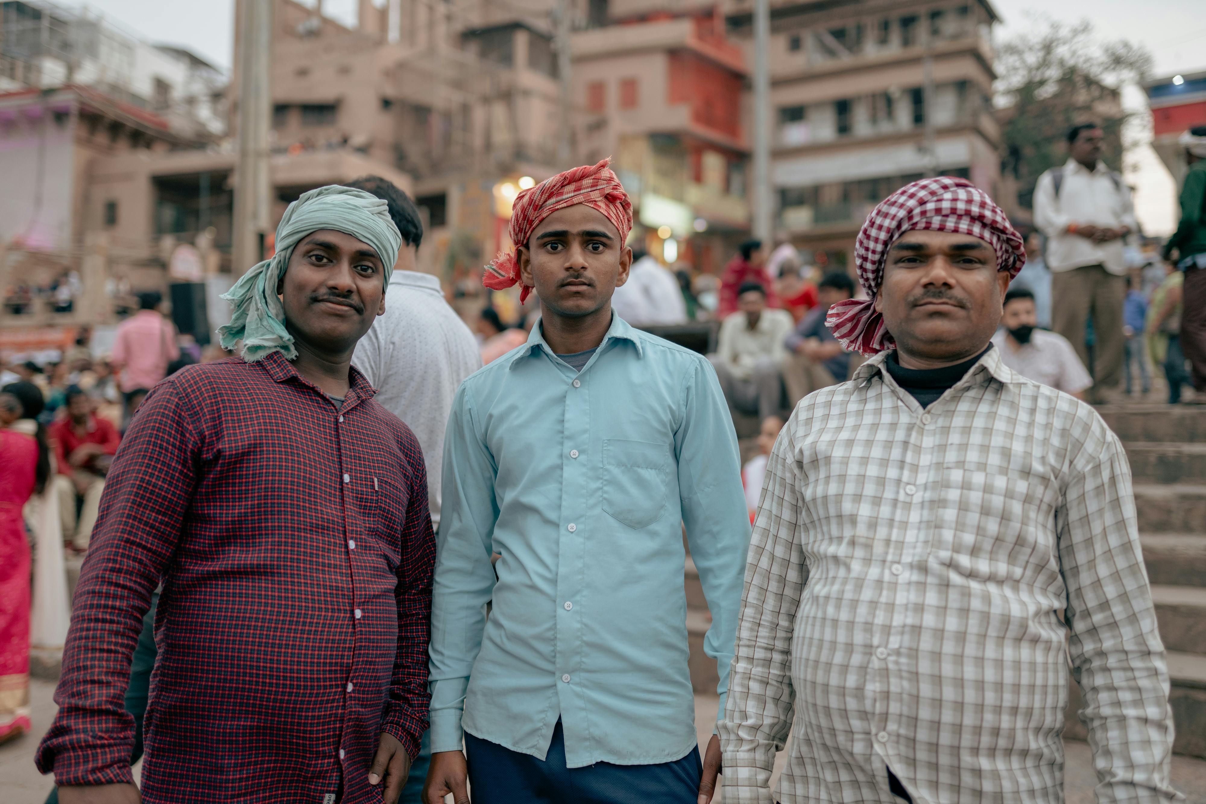Three men wearing traditional headscarves stand at Varanasi ghats, showcasing cultural attire and vibrant local life.